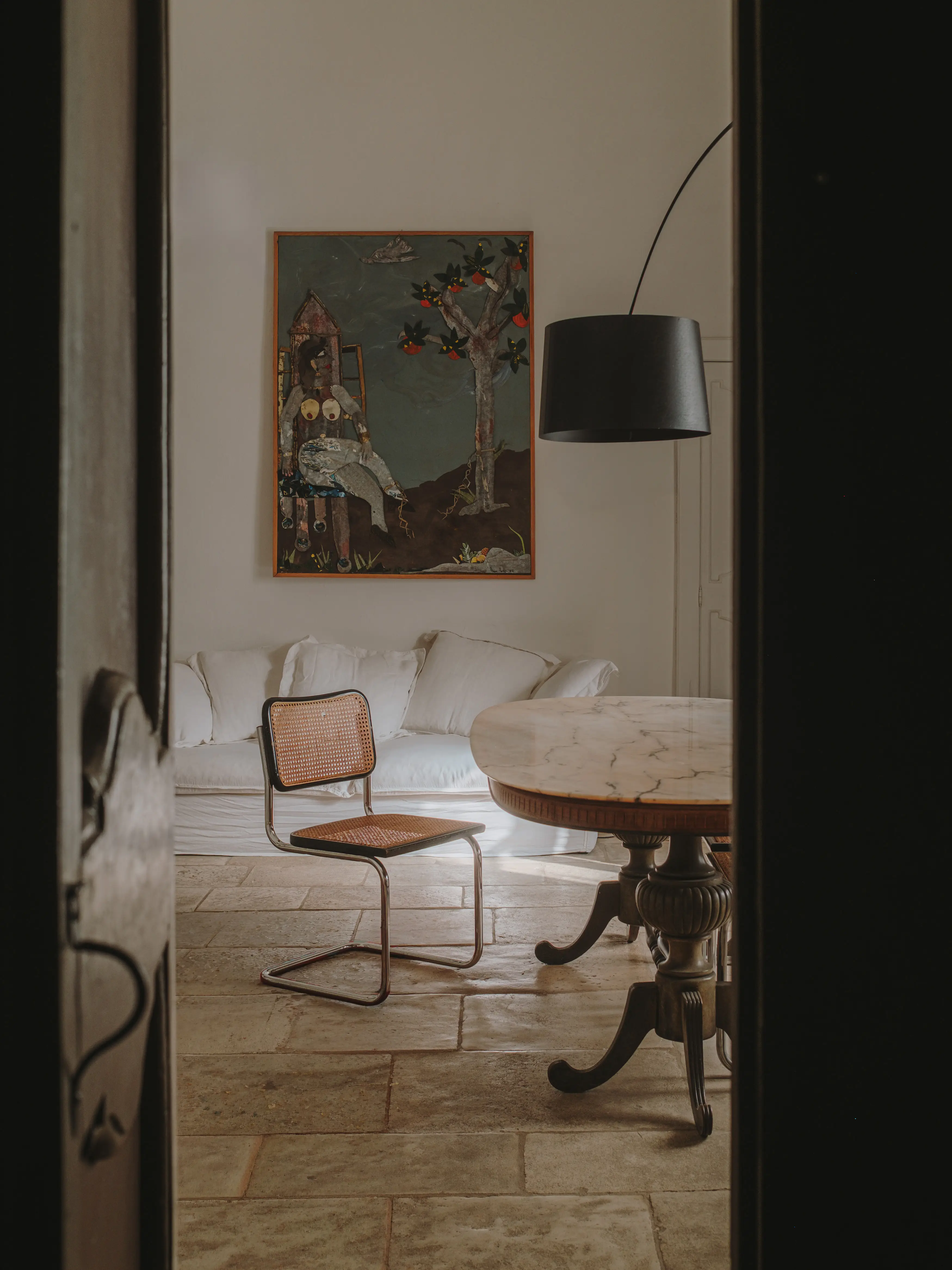 Detalle de un interior minimalista con silla de rejilla, mesa de mármol y un cuadro llamativo, en tonos cálidos y naturales.