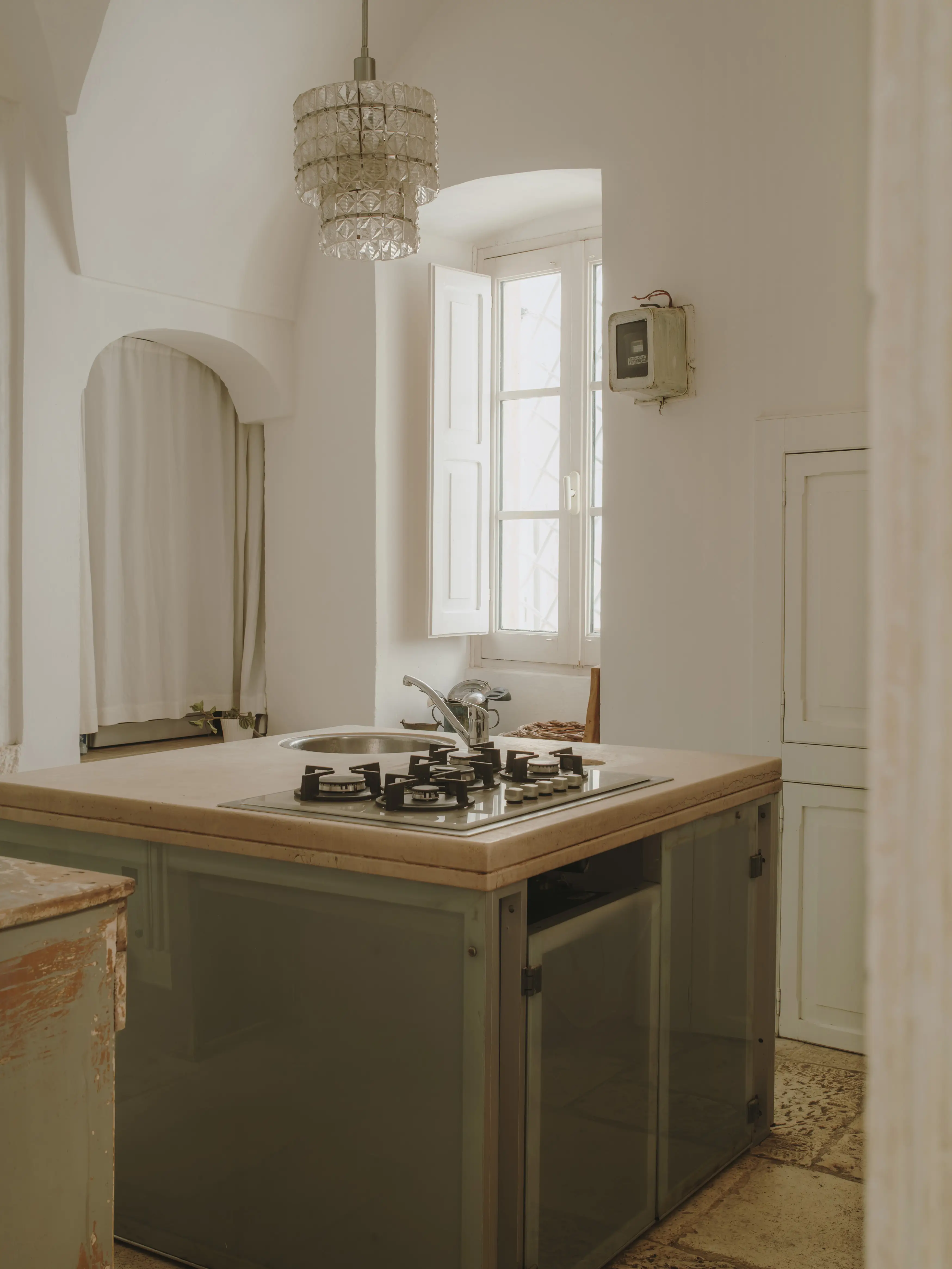 Cocina luminosa con isla central equipada, fregadero y fogones de gas, bajo una elegante lámpara de araña y junto a una ventana con persianas.