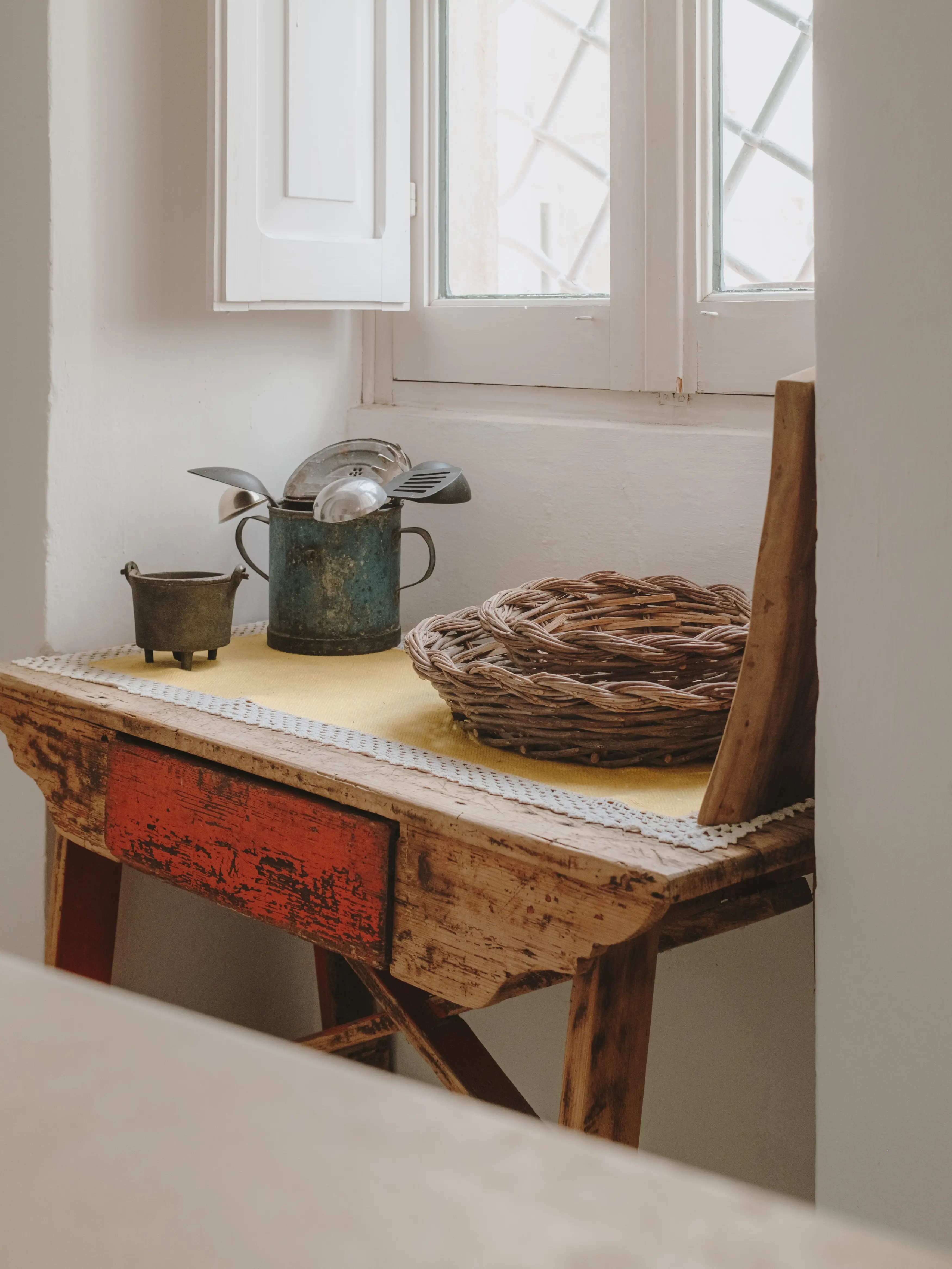 Rincón rústico con mesa de madera, utensilios de cocina en jarra vintage, cesto de mimbre y ventana luminosa.