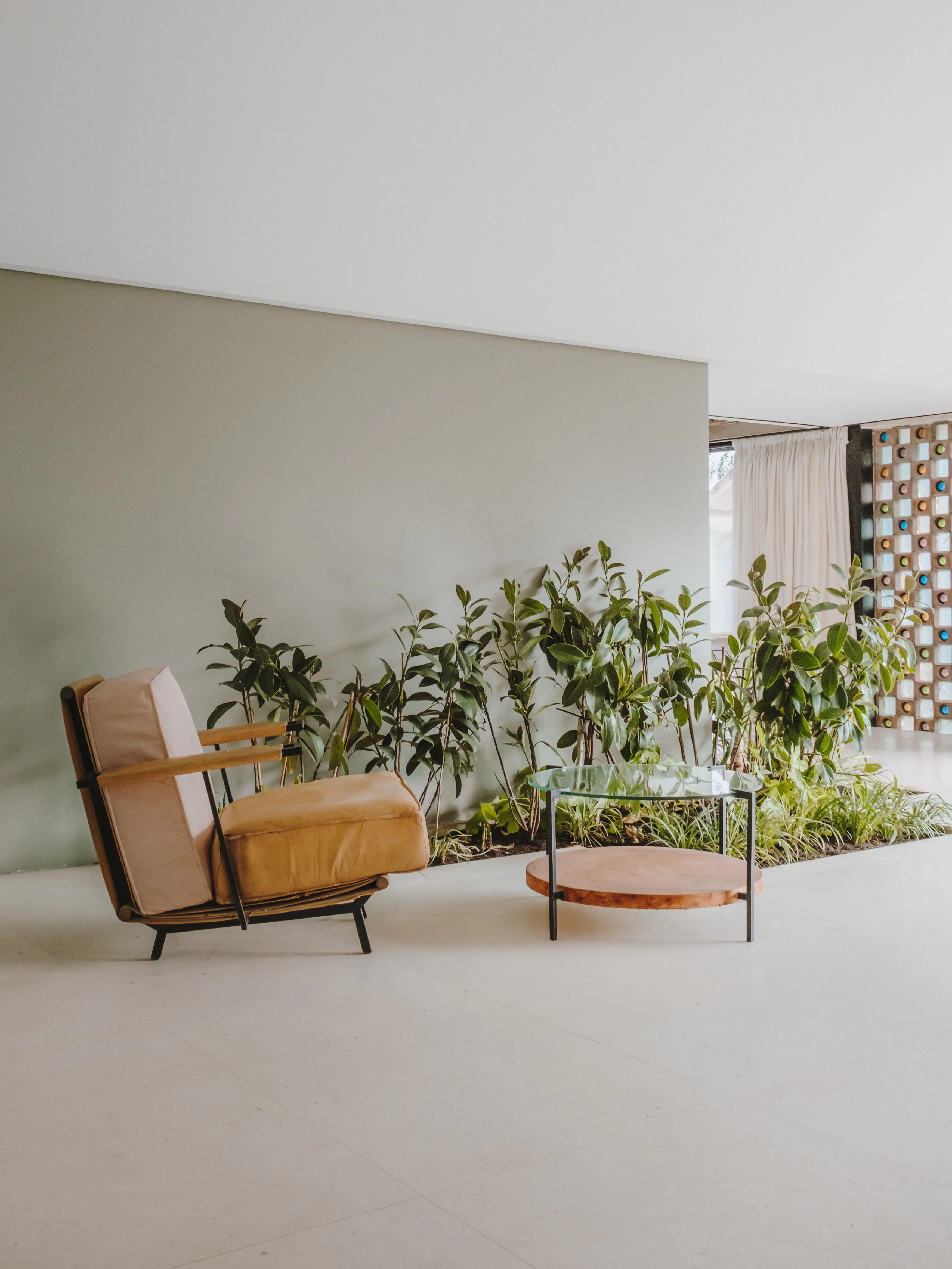 Acogedor rincón de salón interior con sillón de madera y cojines beige, mesa auxiliar redonda y una exuberante jardinera integrada.