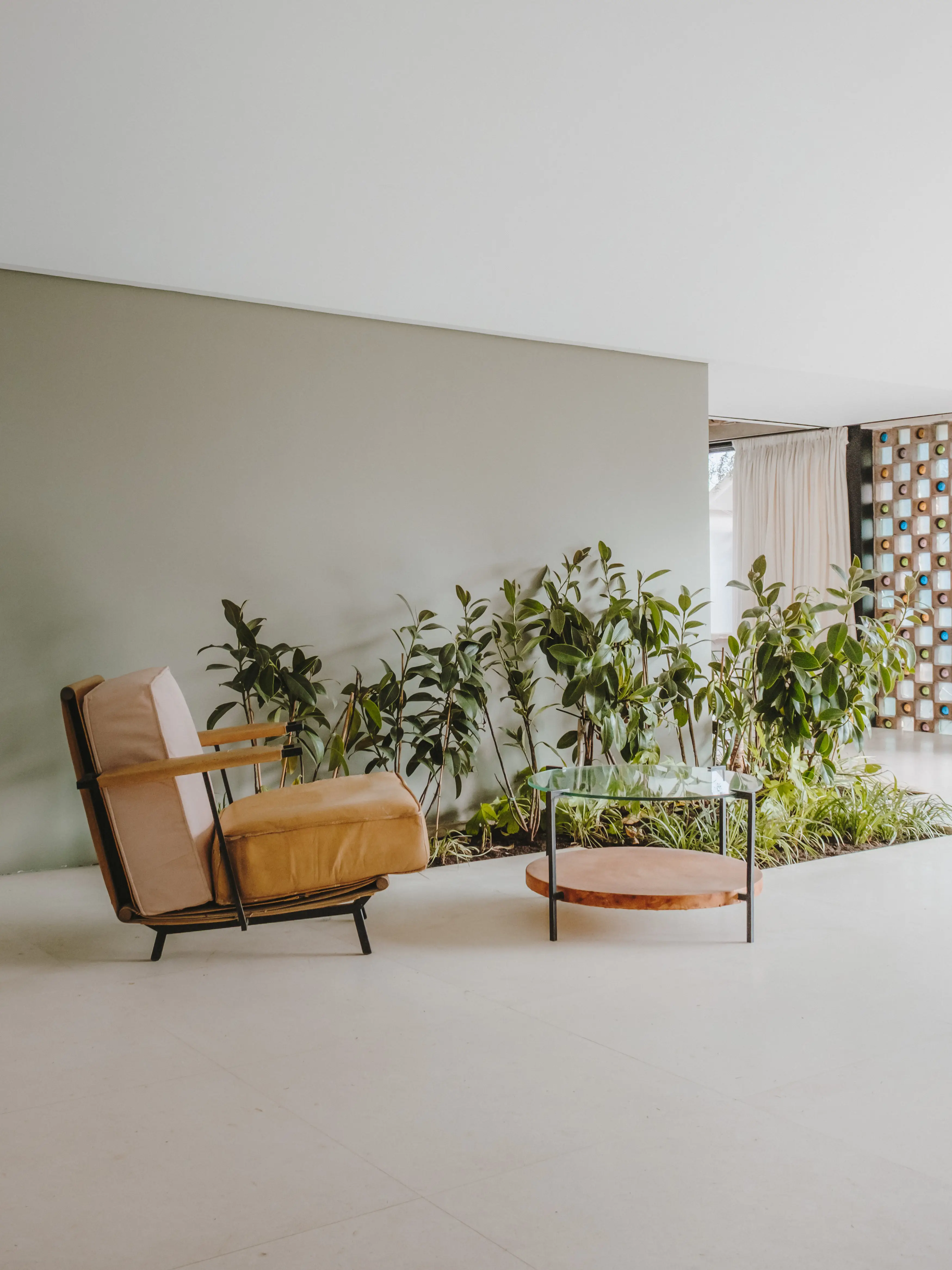 Acogedor rincón de salón interior con sillón de madera y cojines beige, mesa auxiliar redonda y una exuberante jardinera integrada.