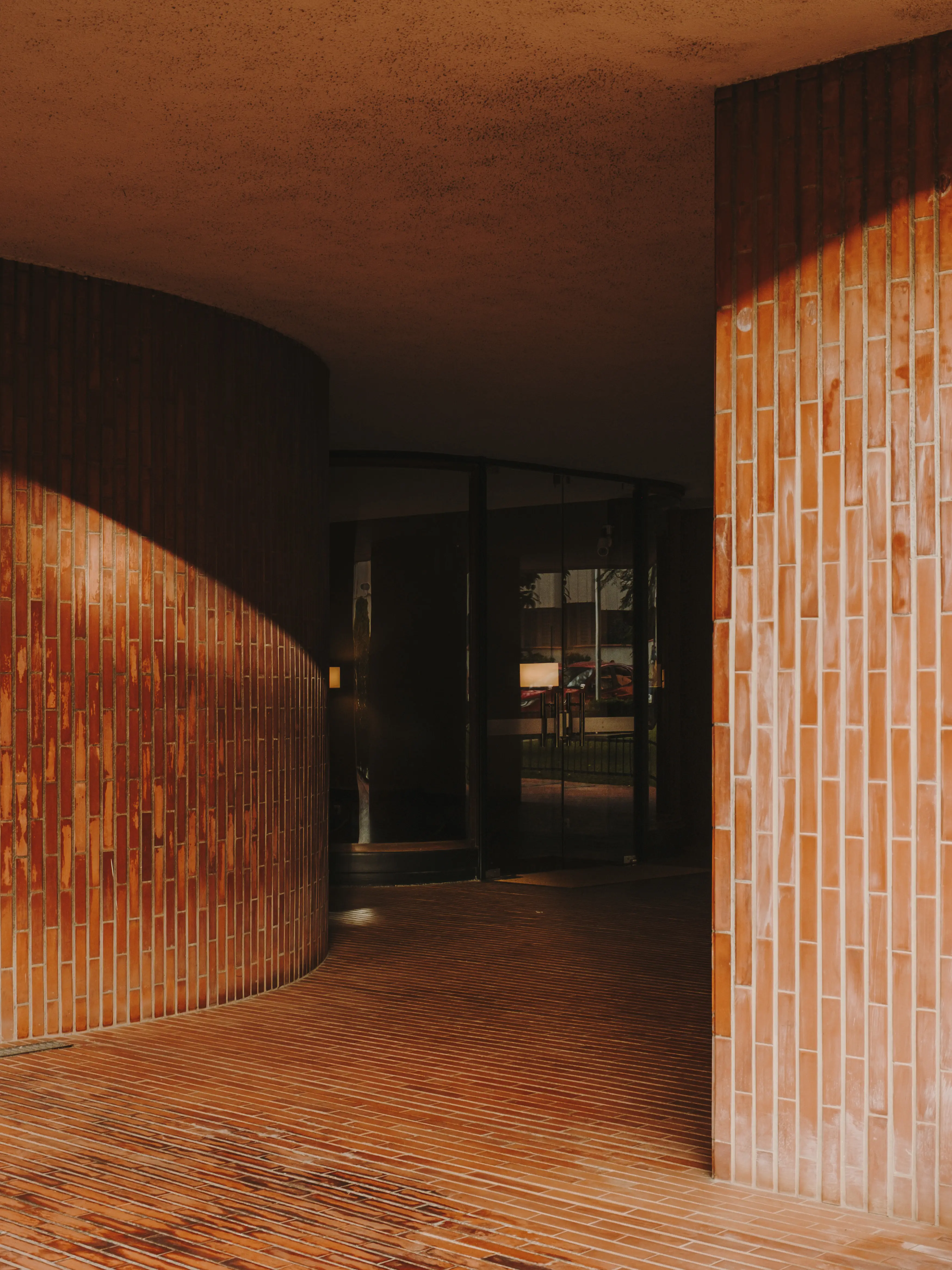 Pasillo arquitectónico con paredes y suelo de ladrillo curvado y recto, iluminado por el sol, con entrada de cristal oscuro al fondo.